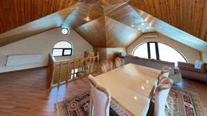 A spacious attic room with a wooden sloped ceiling, a dining table with chairs, a sofa, and a large window. - Spacious 7-bedroom house in peaceful Baku with AC (Baku)