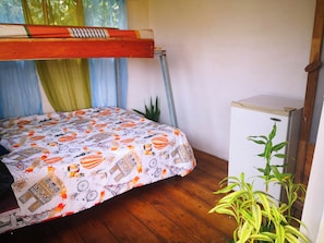 Quarto com cama de solteiro, pequeno frigorífico, planta em vaso e janela com cortinas.