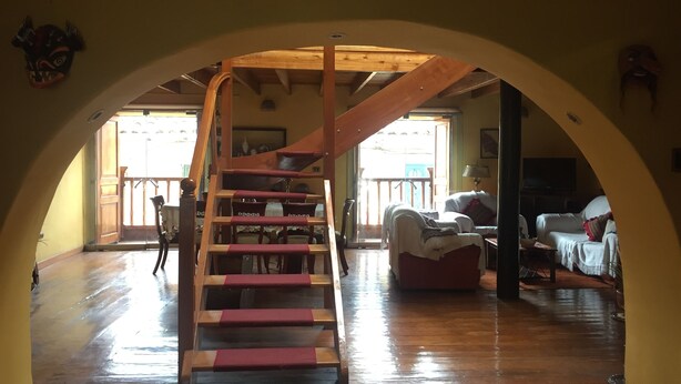 Una escalera de madera con escalones rojos que sube a una zona superior, una sala de estar con sofá y sillones, y vistas al exterior a través de un ventanal.