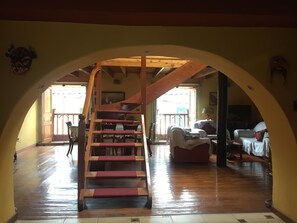 Eine Holztreppe mit roten Stufen führt zu einem erhöhten Bereich, einem Wohnzimmer mit Sofa und Sesseln, und einem Blick ins Freie durch eine Glastür.