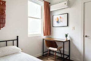Ein Schlafzimmer mit Bett, Schreibtisch mit Stuhl, Fenster mit Vorhängen, einer Klimaanlage und einem gerahmten Bild an der Wand.