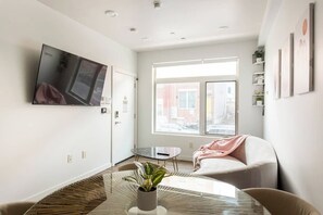 Ein modernes Wohnzimmer mit Fernseher, einem runden Glastisch mit Pflanze, einer Couch und einem Fenster mit Blick auf Gebäude.