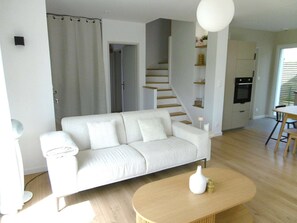 Ein modernes Wohnzimmer mit einem weißen Sofa, einem Holztisch, einer Treppe und einem Küchenbereich.