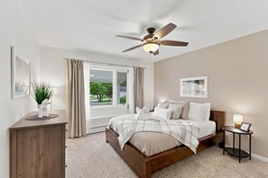 Un dormitorio con cama, ventilador de techo, ventana con cortinas, mesita de noche con lámpara y un cuadro enmarcado en la pared.