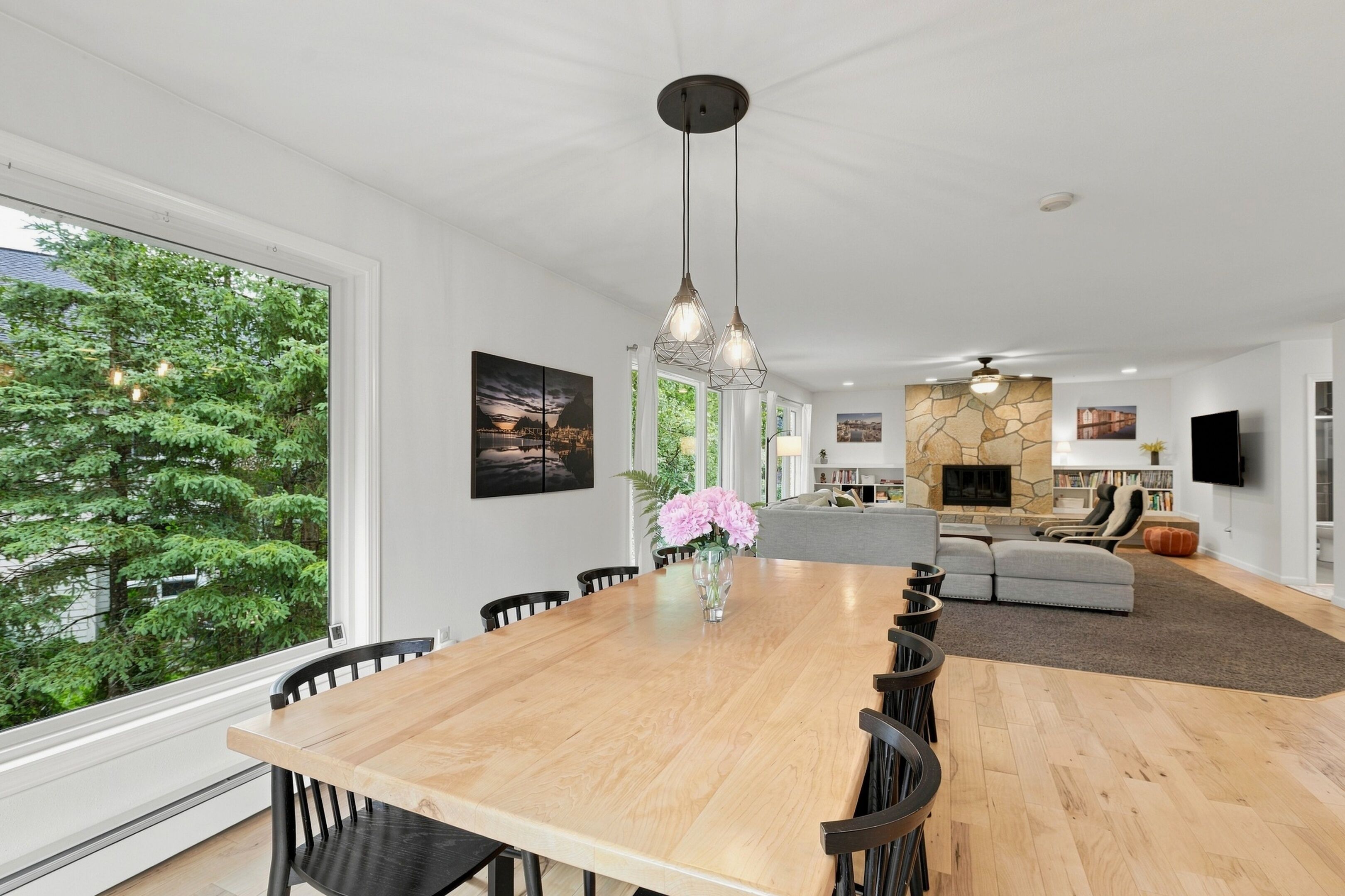 A spacious dining area with a wooden table and chairs, a large window with a view of trees, and a stone fireplace in the background.