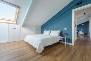 Ein Schlafzimmer mit schrägen Wänden, einem Bett, einem Nachttisch, einer roten Lampe und Blick auf den angrenzenden Raum.