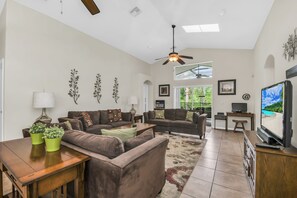 A spacious living room with a ceiling fan, a flat-screen TV, and a large window offering a view of greenery. - Disney World Retreat Oasis & Private Pool (Kissimmee)