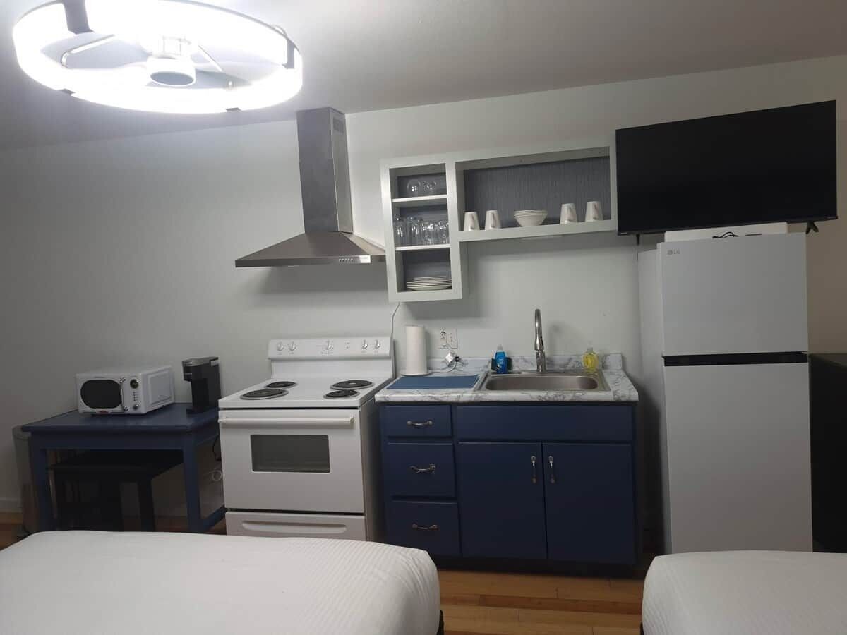 A compact hotel room with a kitchenette, a bed, a TV, and a ceiling light.