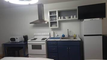 A compact hotel room with a kitchenette, a bed, a TV, and a ceiling light.