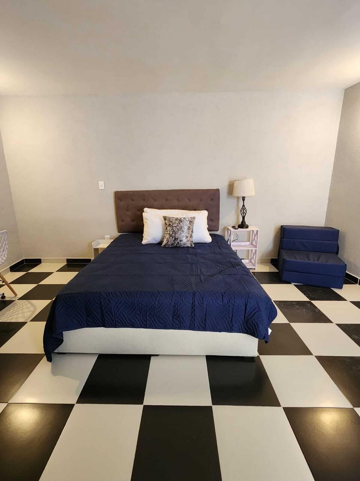 A hotel room with a checkered floor, a bed with a dark blue bedspread, a headboard, a nightstand with a lamp, and a blue bench.