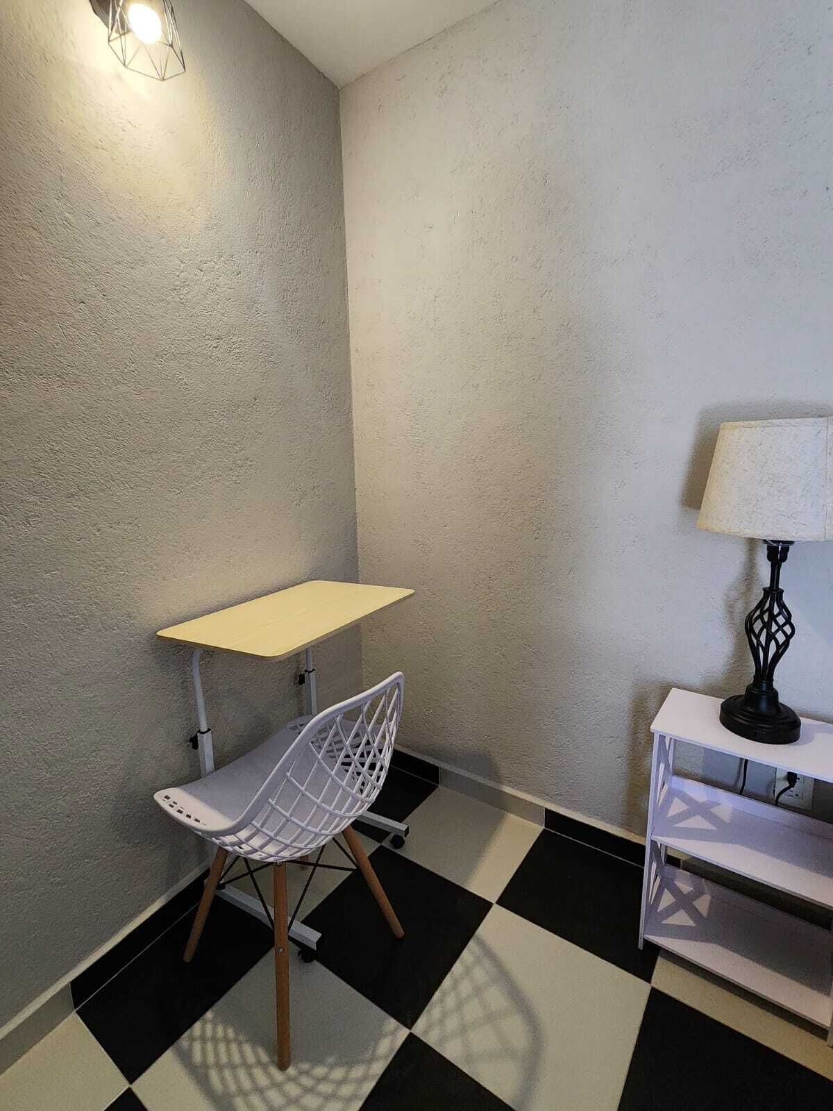 Una habitación pequeña con piso a cuadros, una silla blanca, una mesita y una lámpara sobre una mesita auxiliar.