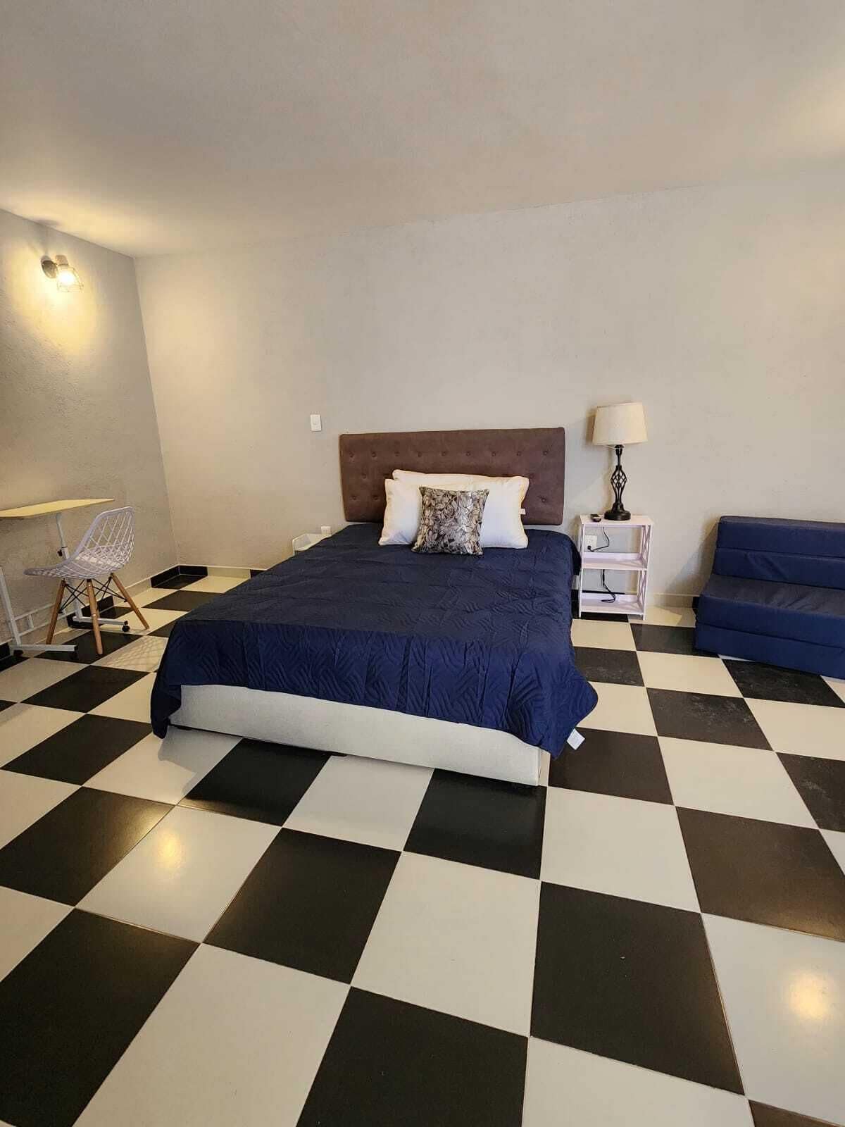 A bedroom with a checkered floor, a bed with a dark blue bedspread, a white lamp on a bedside table, and a blue sofa.