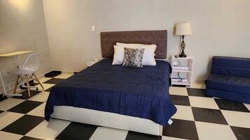 A bedroom with a checkered floor, a bed with a dark blue bedspread, a white lamp on a bedside table, and a blue sofa.