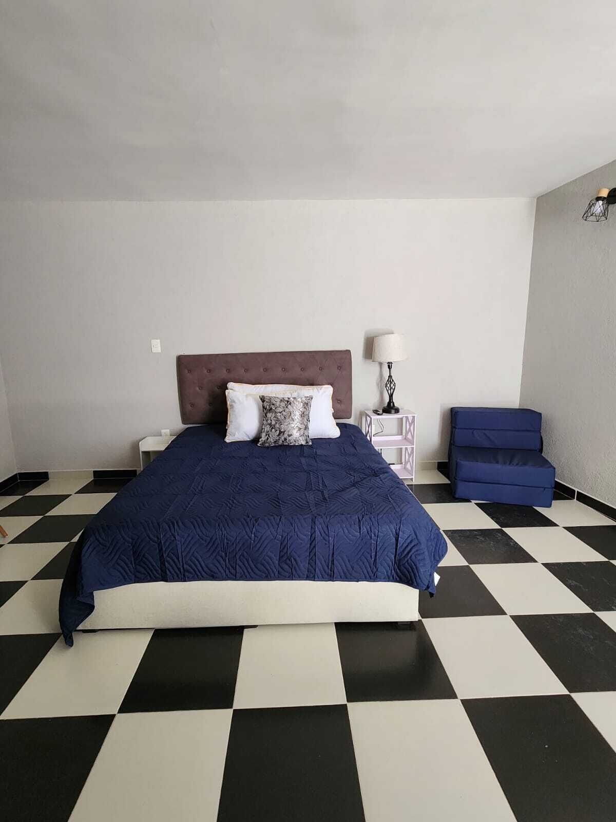 Un dormitorio con piso a cuadros, una cama con cubrecama azul oscuro, cabecera marrón y una mesita de noche con lámpara.
