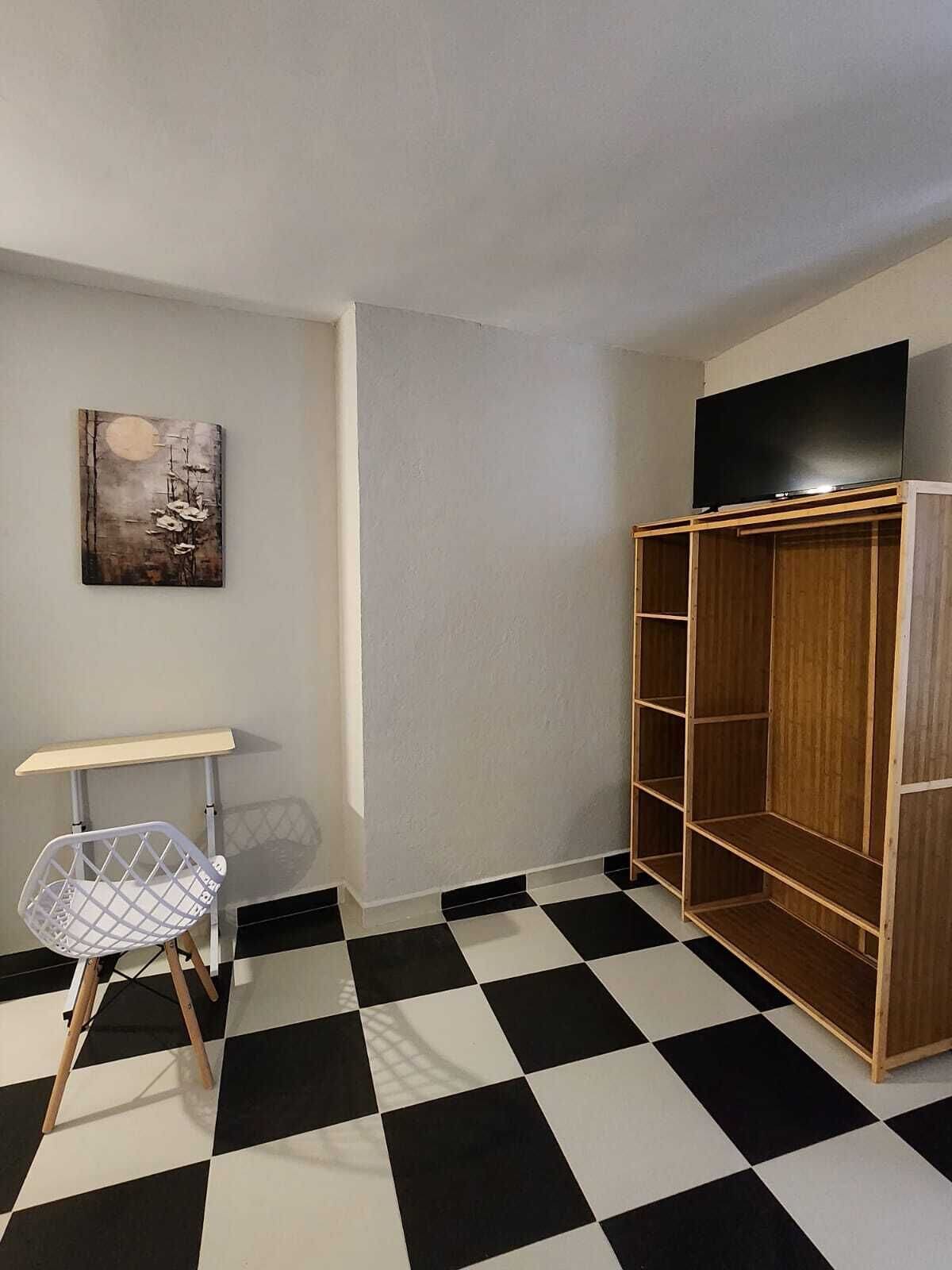 Una habitación con suelo a cuadros, una silla blanca, un armario de madera, un televisor encima y un cuadro en la pared.
