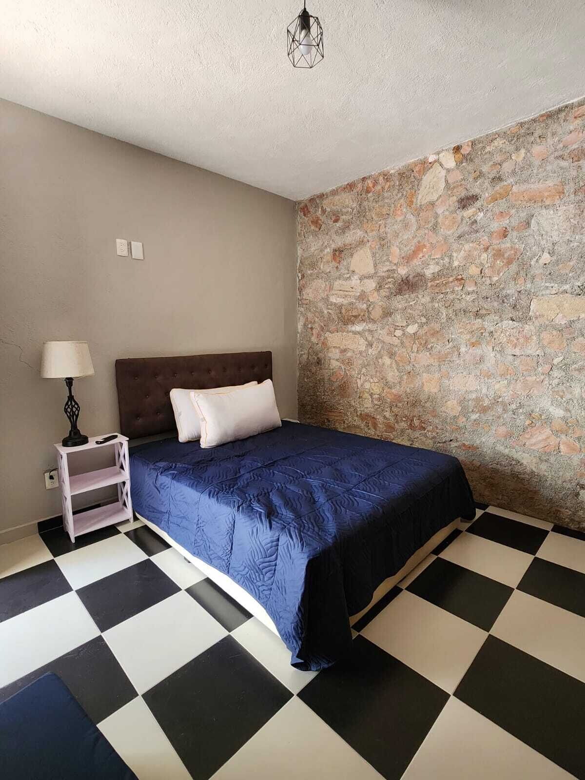 A bedroom with a checkered floor, a bed with a blue bedspread, a nightstand with a lamp, and a stone wall.