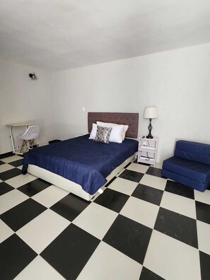 Ein Schlafzimmer mit Schachbrettfußboden, einem Bett mit blauer Bettdecke, einem weißen Nachttisch, einer Lampe und einem blauen Sofa.