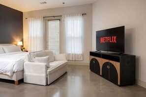 A hotel room with a bed, a white sofa, a TV on a wooden cabinet, and a window with curtains. - Stylish Modern Studio | KING & FULL BED + Pool&Gym (Irving)