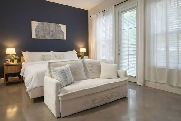 A bedroom with a bed, a white sofa, a nightstand with a lamp, and a wall-mounted artwork. - Stylish Modern Studio | KING & FULL BED + Pool&Gym (Irving)