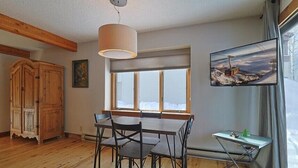 Een eetgedeelte met een tafel en stoelen, een houten kast, een televisie aan de muur, en een raam met uitzicht.