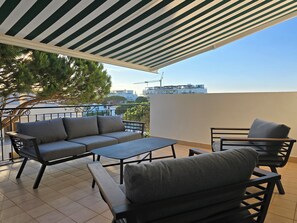 A balcony with a striped awning, a grey sofa, a small table, and a chair. - Falésia Townhouse - Walk to Beach - 3 Terraces (Olhos de Água)