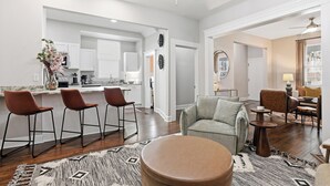 Ein modernes Wohnzimmer mit einer Couch, einem Hocker und Barhocker. Im Hintergrund ist eine Küche mit weißen Schränken und einem Essbereich erkennbar.
