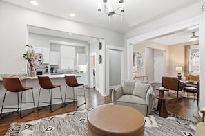 A modern living room with a sofa, ottoman, and bar stools. A kitchen is visible in the background with white cabinets and a dining area. - Savanah Home, Walkable Downtown, 30+ Days (Savannah)