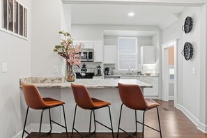 A modern kitchen with white cabinets, a marble countertop, and a central island with stools. - Savanah Home, Walkable Downtown, 30+ Days (Savannah)