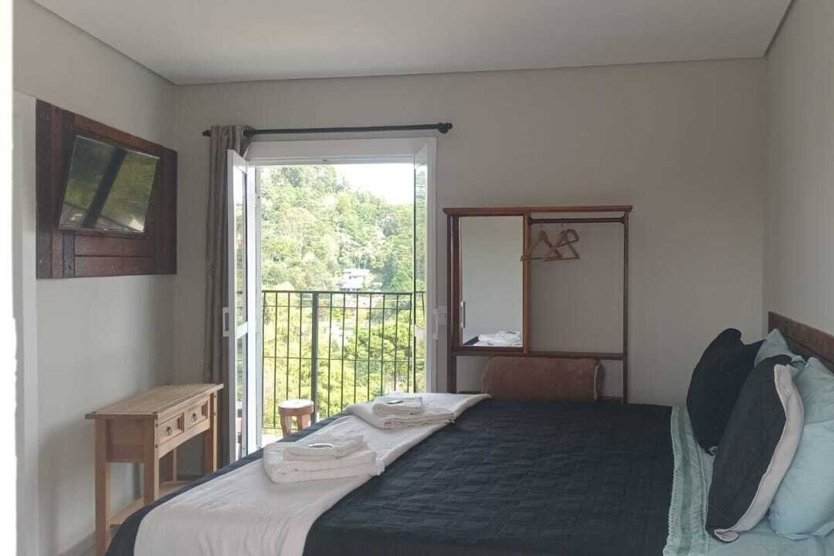 A bedroom with a bed, a bedside table, a mirror, and a window with a view of greenery.