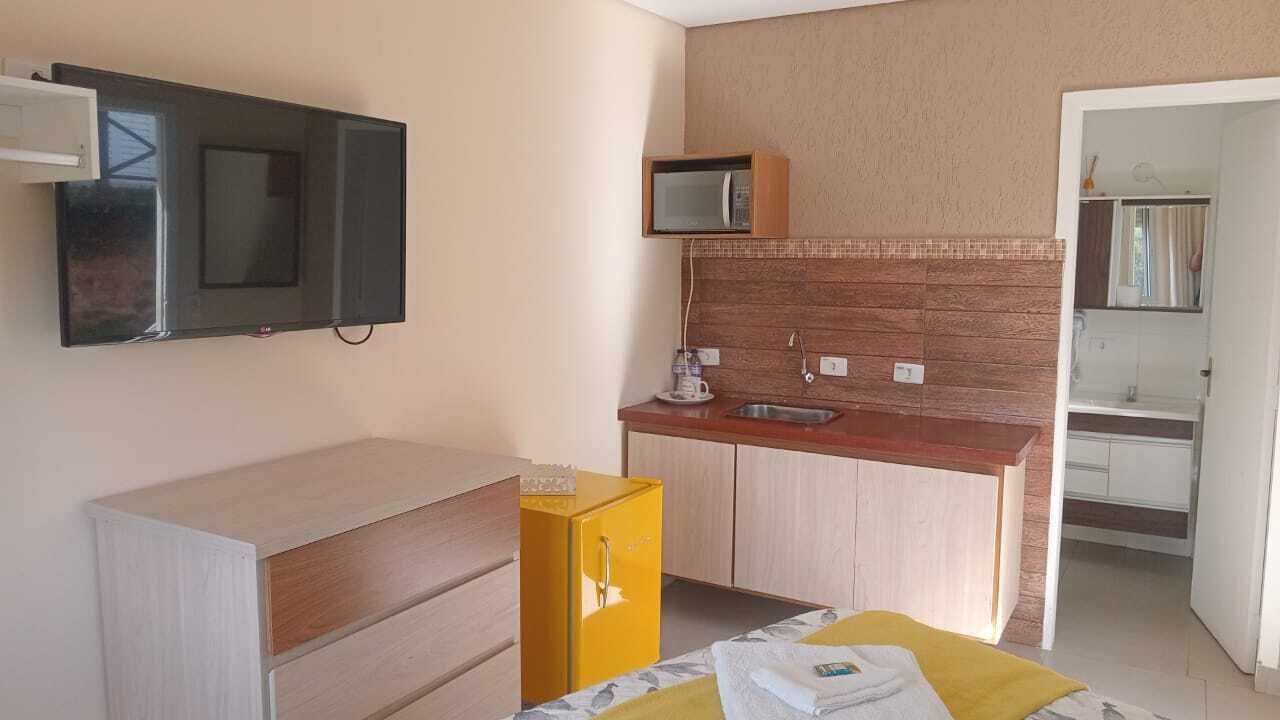 A compact hotel room with a flat-screen TV, a microwave, a small sink, a wooden cabinet, and a bed with a yellow blanket.
