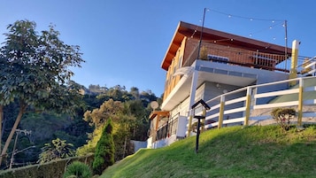 Um edifício moderno de dois andares com varanda, cercado por vegetação exuberante e colinas.