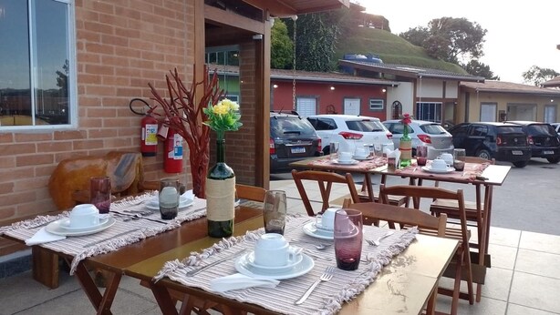 Un área de comedor al aire libre con mesas dispuestas para dos, un edificio de ladrillo, coches estacionados y una colina al fondo.