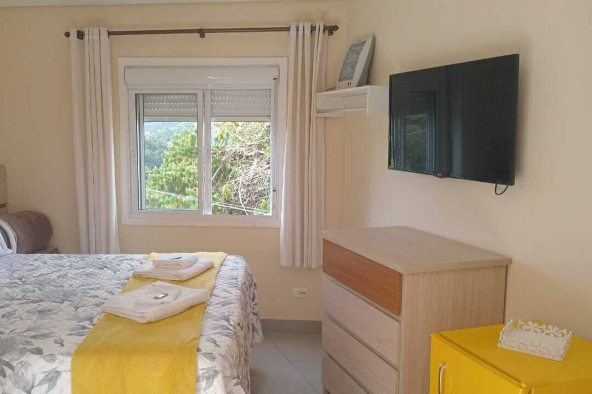 A bedroom with a bed, a window with curtains, a TV mounted on the wall, and a yellow dresser.