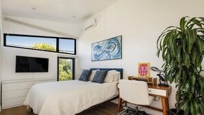 Ein Schlafzimmer mit einem Bett, einem Schreibtisch, einem Stuhl, einer großen Topfpflanze und einem an der Wand montierten Fernseher.