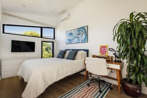 Ein Schlafzimmer mit einem Bett, einem Schreibtisch, einem Stuhl, einer großen Topfpflanze und einem an der Wand montierten Fernseher.