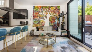 Ein modernes Wohnzimmer mit einer Couch, einem Glastisch, einem farbenfrohen abstrakten Gemälde, einem Fahrrad und einer Glasschiebetür nach draußen.