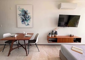 A modern living room with a wooden dining table, white chairs, a mounted TV, and a wall-mounted shelf with decorative items. - Ocean-View King Studio with Private Balcony & Rooftop Pool (Playa del Carmen)