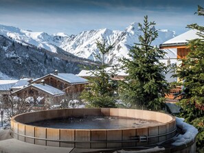 A wooden hot tub surrounded by snow-covered mountains and buildings.