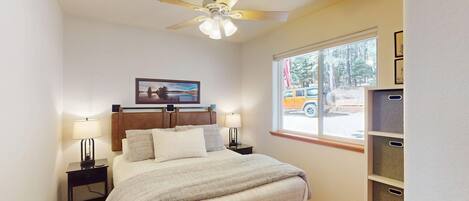 A bedroom with a bed, two bedside tables, a ceiling fan, a window with a view of a vehicle, and a framed picture on the wall.