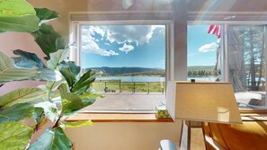 Quarto com uma grande janela com vista para um lago e montanhas, um deck de madeira na parte externa e uma planta no parapeito.