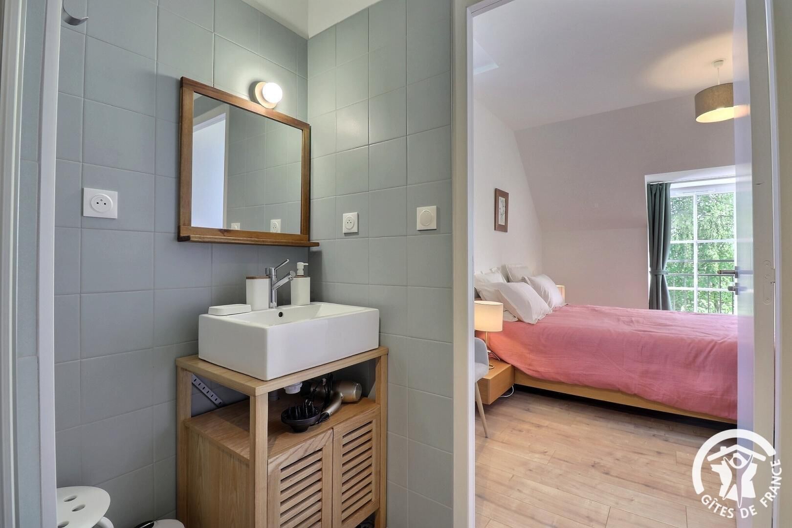 A bedroom with a bed, a bedside table, a window, and a bathroom with a sink and mirror.