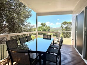 A covered outdoor dining area with a table and chairs, a view of trees and hills, and a metal railing.