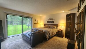 Quarto com cama, sofá, mesa de cabeceira, luminária, espelho e porta de vidro corrediça para um jardim.