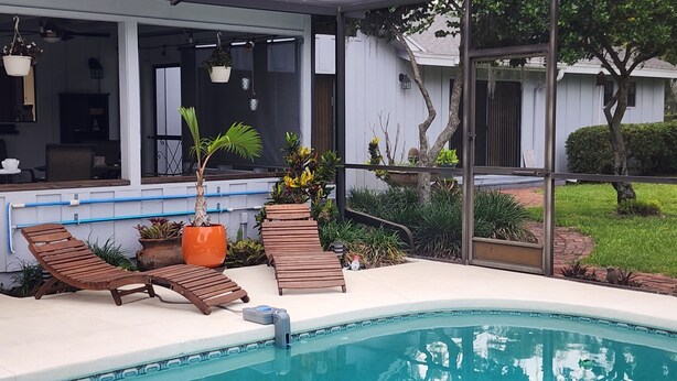 Piscina com espreguiçadeiras, planta em vaso e vista para uma casa com uma janela grande.