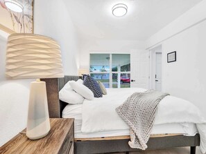 A bedroom with a bed, a bedside lamp, a window with blinds, and a door. - Cool Blue - Designer 3BR Retreat (Fort Lauderdale)