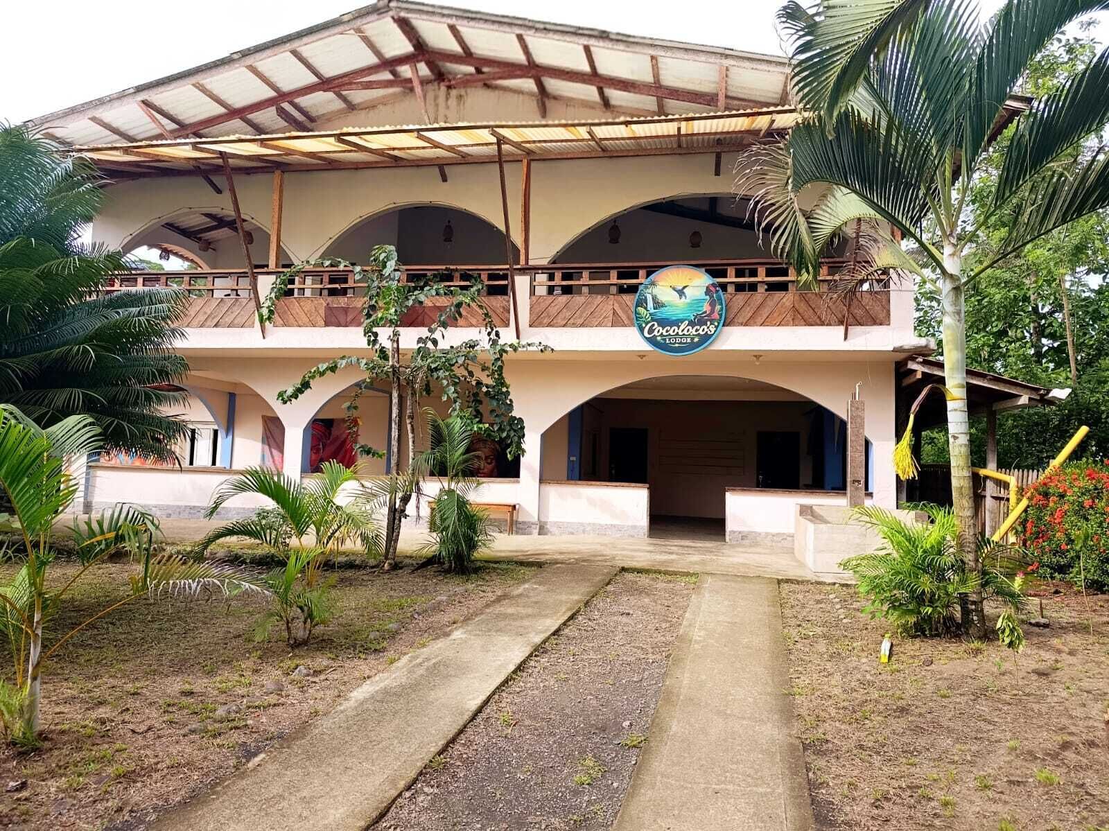 Un edificio de dos pisos con un porche, un letrero que dice 'Cocotoco's Lodore' y un jardín con palmeras.