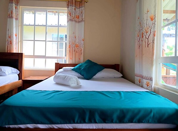 A single bed with a teal bedspread, white pillows, and a rolled white towel. There is a window with a view of the outside, and curtains with a floral pattern.