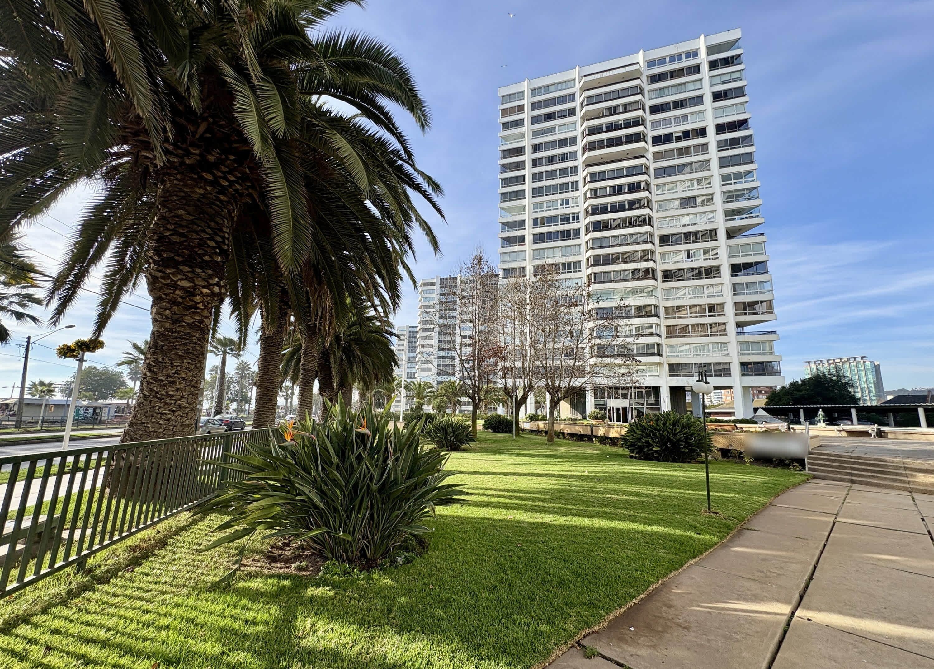 Un edificio moderno con techo plano y varios balcones, rodeado de césped cuidado y palmeras.
