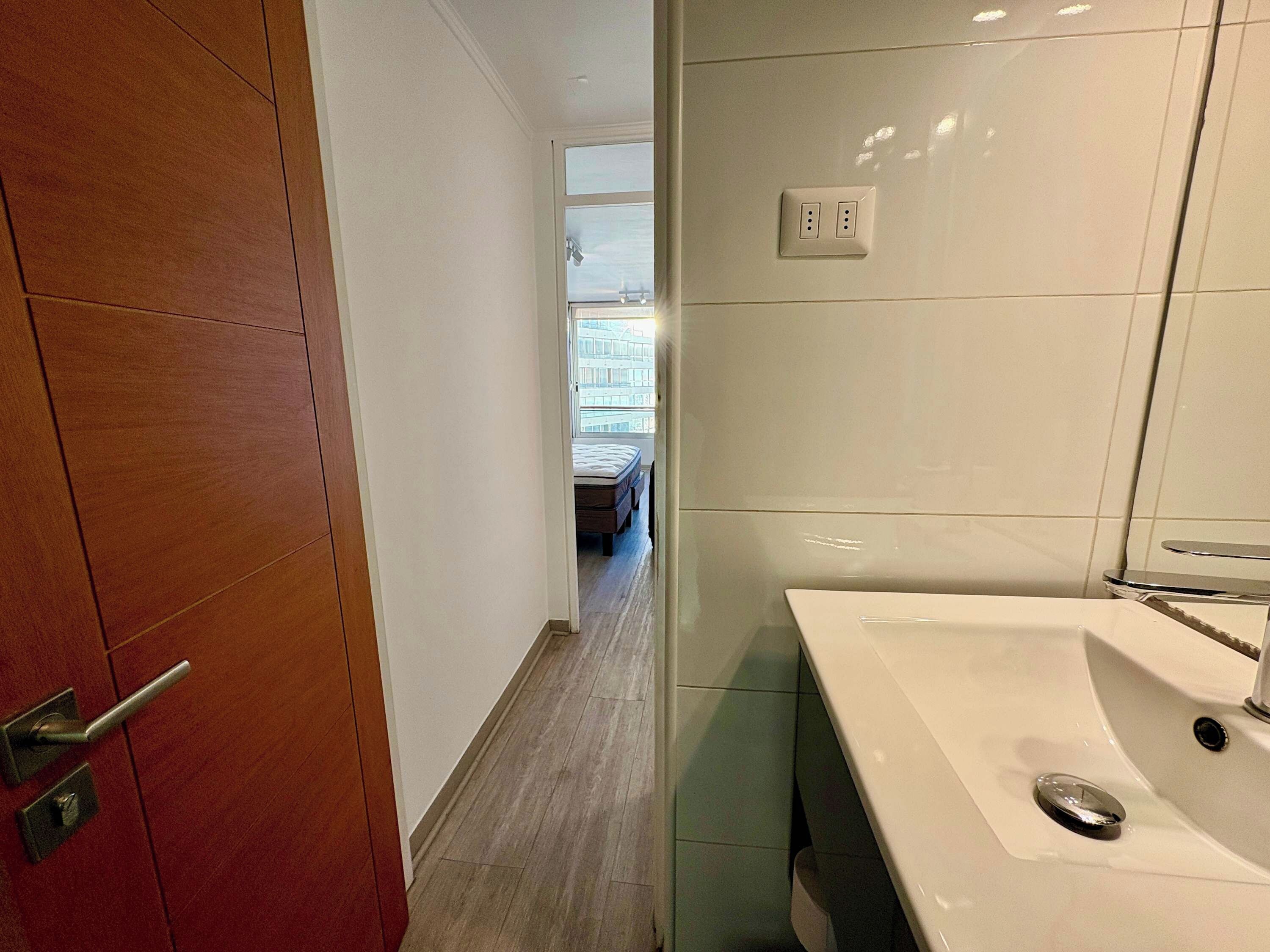 A hotel room with a wooden door, a white wall, a large window, and a bathroom with a sink and mirror.