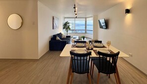 A dining area with a table set for four, a sofa, a TV mounted on the wall, and a large window with a view.